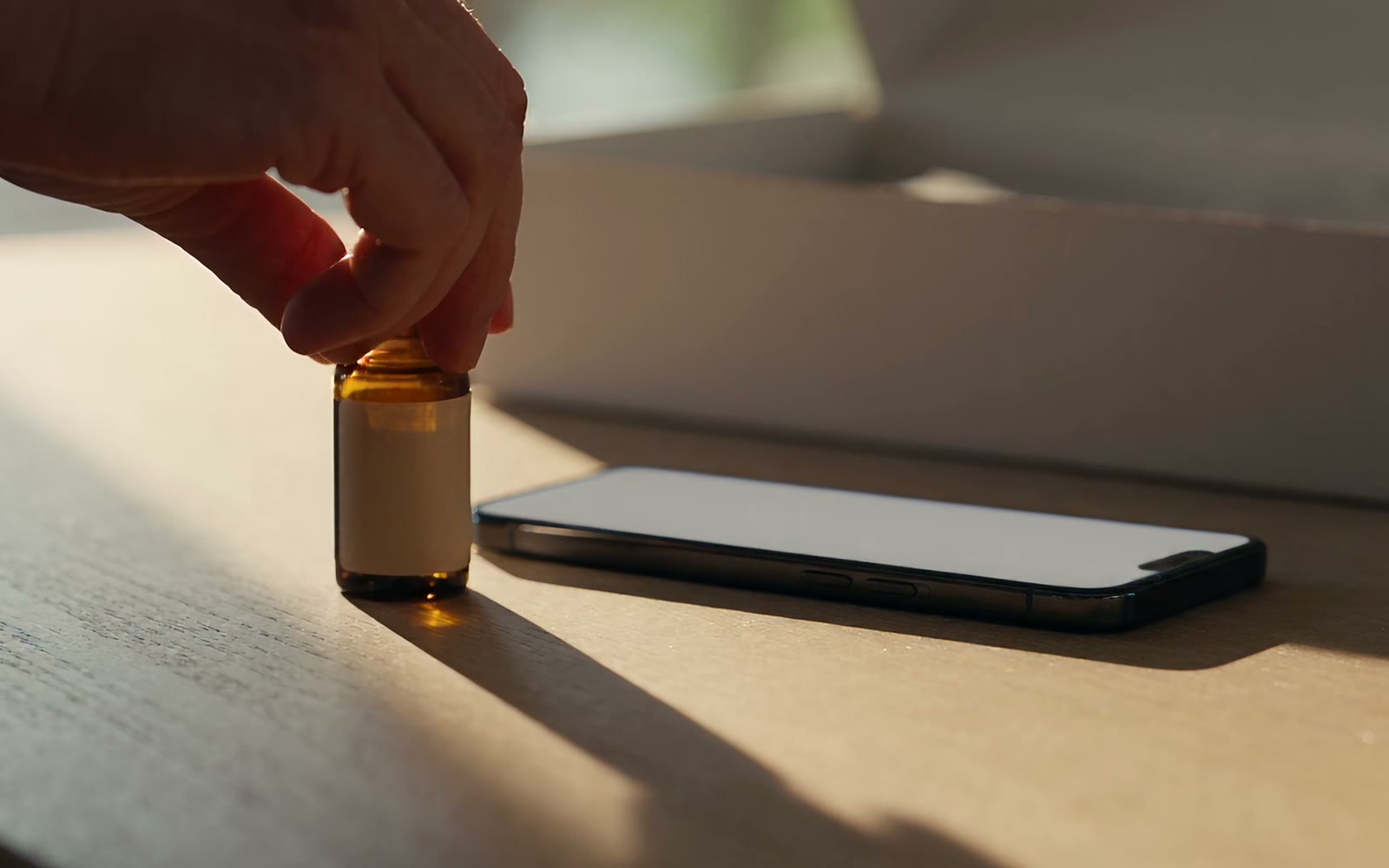 A hand placing a single amber supplement bottle beside a phone on a sunlit oak table.