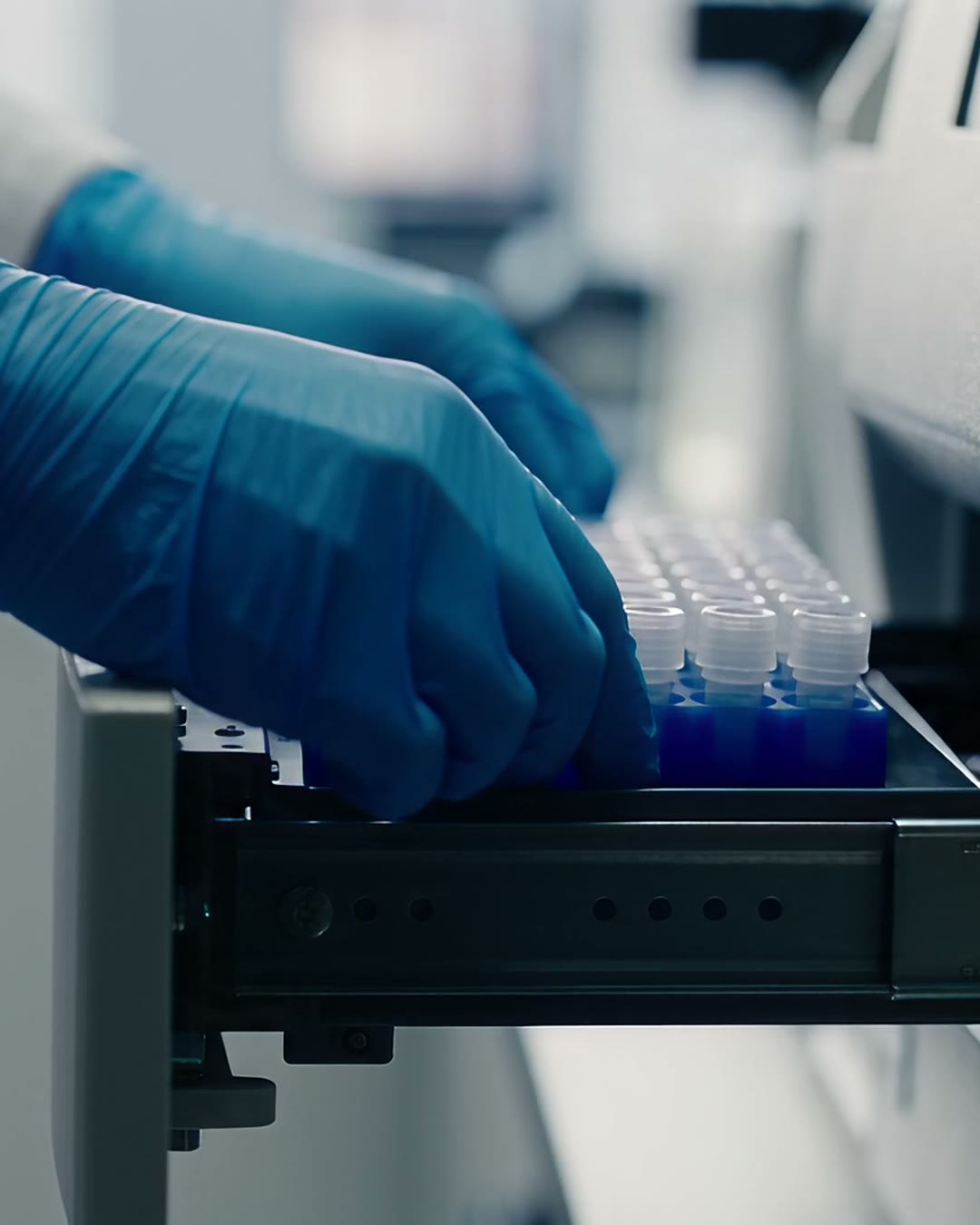 A laboratory technician in blue gloves loading a rack of capped sample tubes into an analyser.